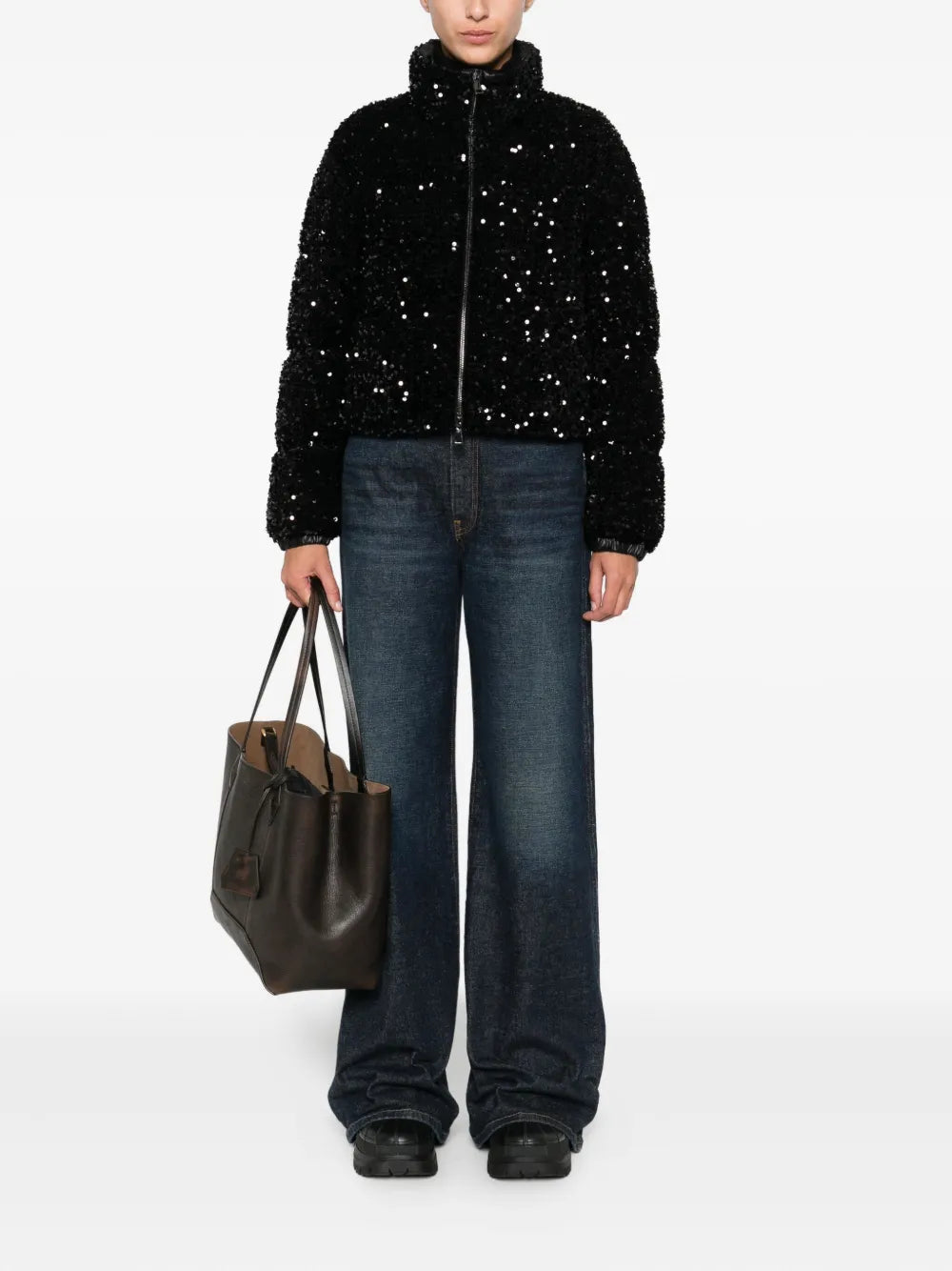 Woman wearing a black Moncler Louvi Puffer Jacket with sequin finish, high collar, and cropped fit, paired with dark wide-leg jeans, black shoes, and carrying a large brown leather tote bag, standing against a white background.